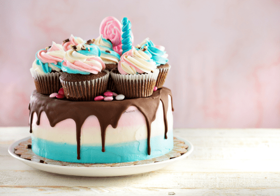 A pink and blue cake with chocolate icing dripping over the edge decorated with colourful cupcakes on top.